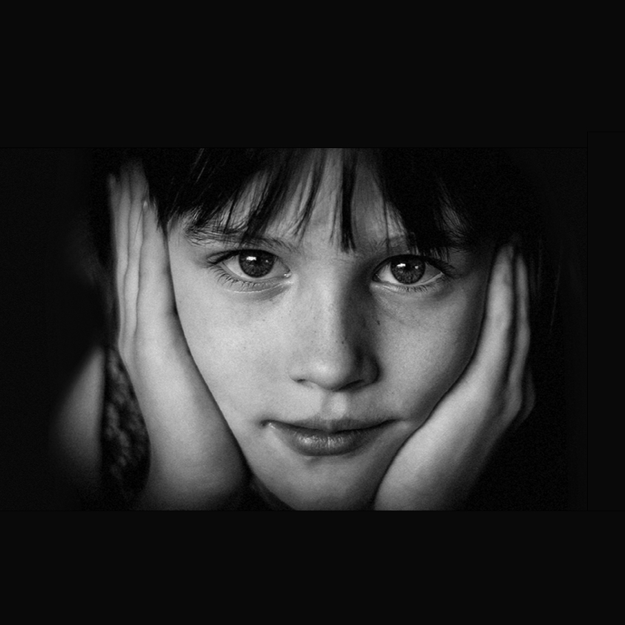 Portræt i sort-hvid af et barn, der hviler ansigtet i hænderne og ser direkte ind i kameraet med et roligt, eftertænksomt udtryk.
